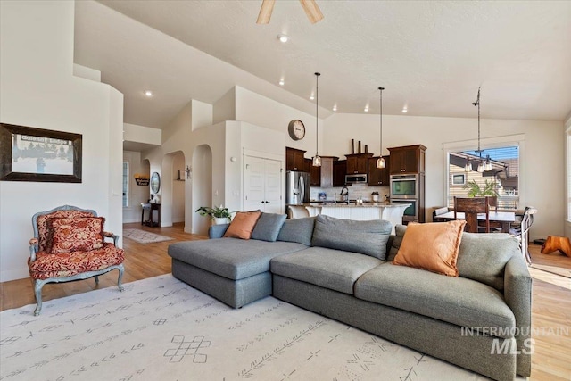 living room with light wood-type flooring, arched walkways, high vaulted ceiling, and ceiling fan with notable chandelier