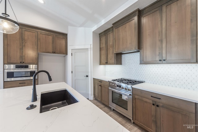 kitchen with stainless steel appliances, hanging light fixtures, a sink, light stone countertops, and wall chimney exhaust hood