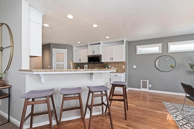 kitchen featuring white cabinets, light wood-type flooring, decorative backsplash, stainless steel microwave, and a kitchen bar