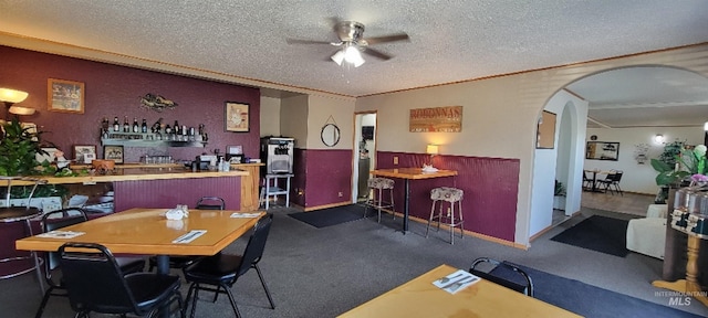 dining area featuring a textured ceiling, dark colored carpet, ceiling fan, and indoor bar