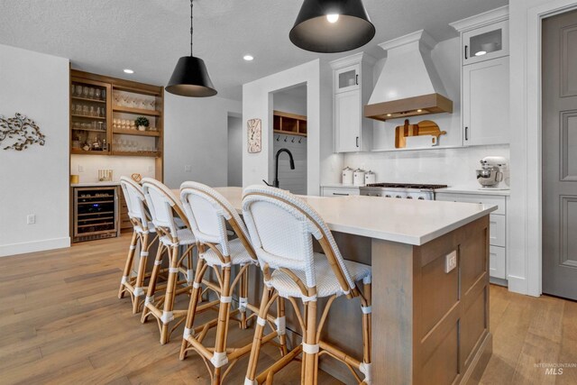 kitchen with wine cooler, custom exhaust hood, open shelves, light countertops, and an island with sink