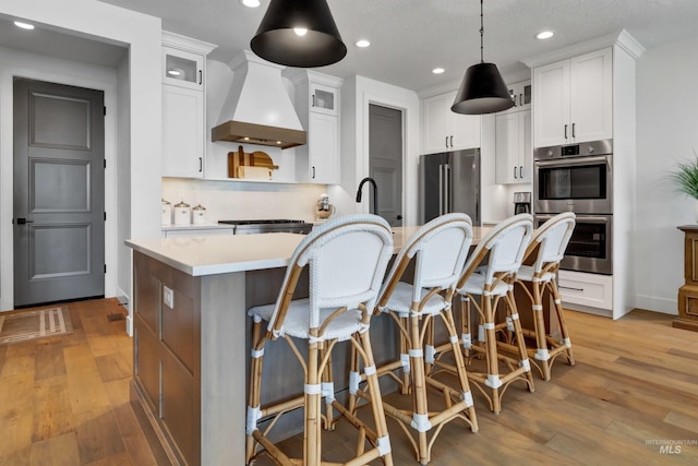 kitchen with appliances with stainless steel finishes, custom exhaust hood, a center island with sink, light countertops, and light wood-style flooring