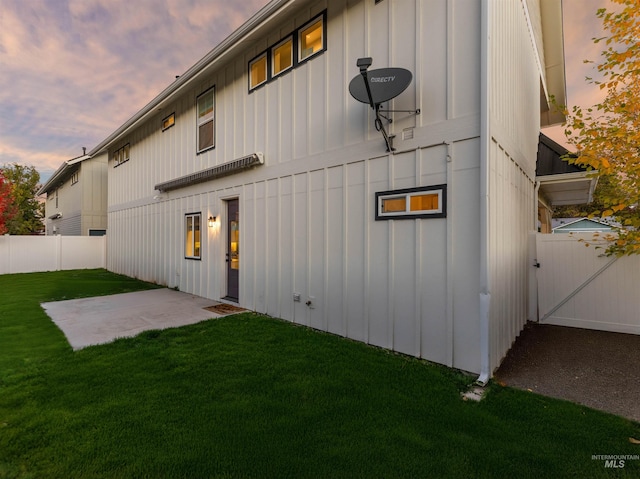 back of house at dusk featuring board and batten siding, a lawn, a patio, fence, and a gate