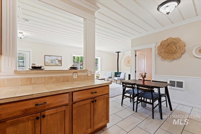 kitchen with brown cabinetry, decorative columns, tile counters, a wainscoted wall, and light tile patterned floors