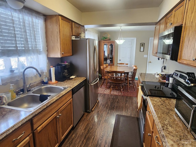 kitchen featuring stainless steel appliances, a sink, brown cabinetry, dark wood finished floors, and decorative light fixtures