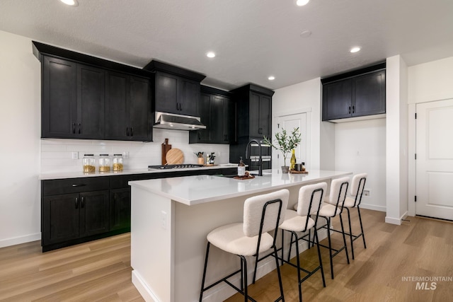 kitchen featuring dark cabinets, decorative backsplash, light wood-style floors, under cabinet range hood, and a center island with sink