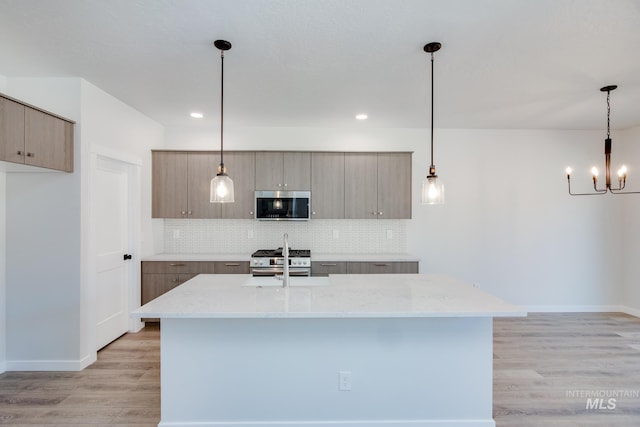 kitchen with tasteful backsplash, appliances with stainless steel finishes, pendant lighting, light stone countertops, and a kitchen island with sink