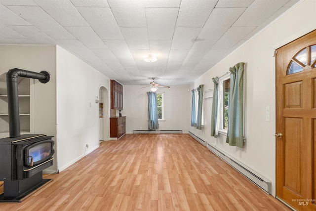 unfurnished living room with a baseboard heating unit, a wood stove, light wood-style flooring, and ceiling fan