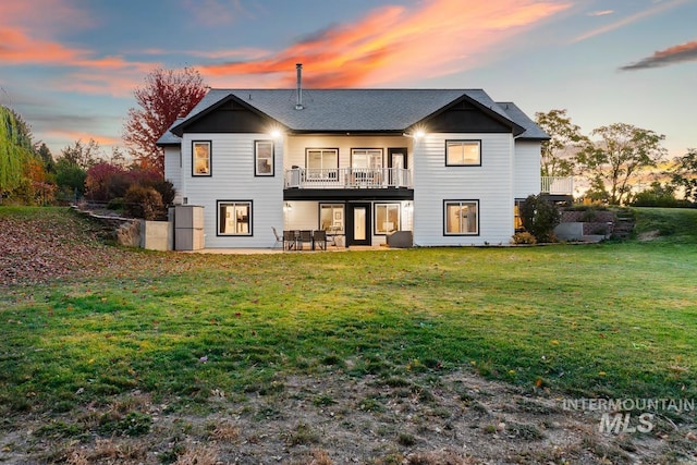 back house at dusk featuring a balcony, a patio area, and a lawn