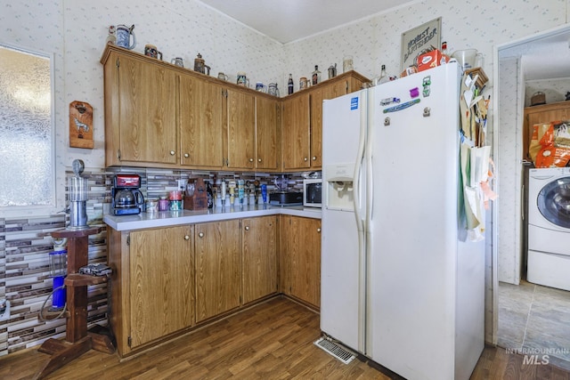 kitchen with wood finished floors, wallpapered walls, white refrigerator with ice dispenser, washer / dryer, and light countertops