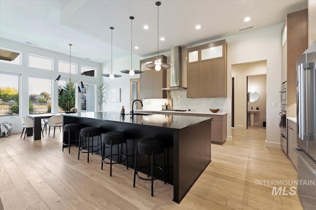 kitchen featuring stainless steel appliances, light hardwood / wood-style flooring, pendant lighting, wall chimney exhaust hood, and a kitchen island with sink