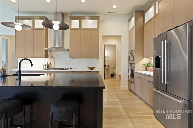 kitchen with a breakfast bar, appliances with stainless steel finishes, wall chimney exhaust hood, decorative light fixtures, and light wood-type flooring