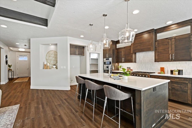 kitchen featuring dark wood-type flooring, premium range hood, decorative light fixtures, dark brown cabinets, and an island with sink