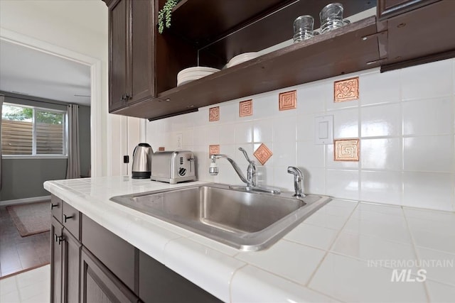 kitchen with a sink, backsplash, dark brown cabinetry, tile countertops, and open shelves