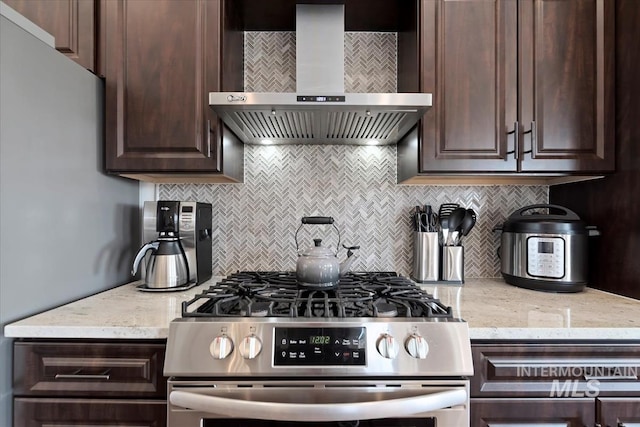 kitchen featuring tasteful backsplash, dark brown cabinets, wall chimney range hood, stainless steel range with gas stovetop, and light stone counters