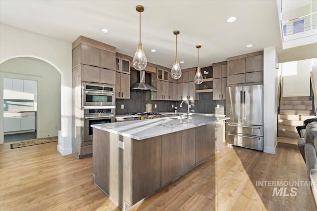 kitchen featuring backsplash, a center island with sink, wall chimney range hood, appliances with stainless steel finishes, and light hardwood / wood-style floors