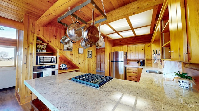 kitchen with wooden walls, a peninsula, a sink, appliances with stainless steel finishes, and beamed ceiling