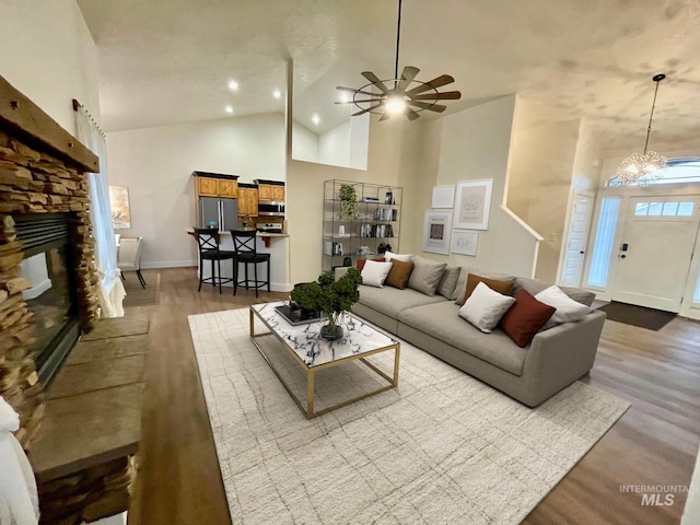 living room with ceiling fan with notable chandelier, a stone fireplace, light hardwood / wood-style flooring, and high vaulted ceiling