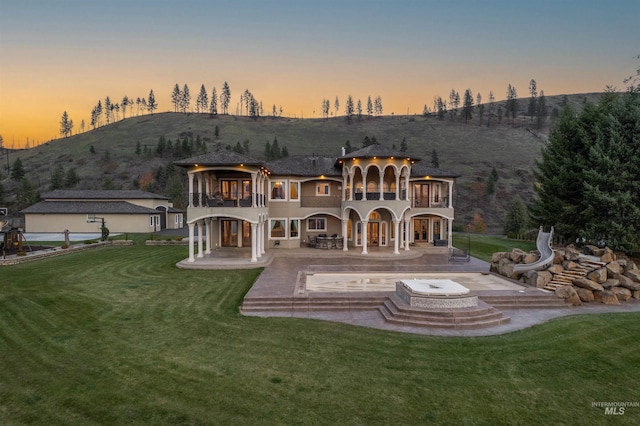 back house at dusk with a lawn, a balcony, a patio, and an outdoor hot tub