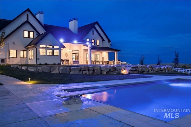 back of house at night with stucco siding, a chimney, a fenced in pool, and a patio