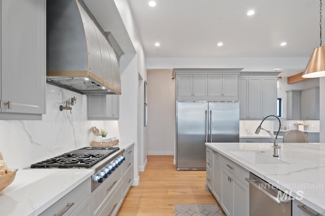 kitchen featuring wall chimney exhaust hood, appliances with stainless steel finishes, light stone countertops, light wood-type flooring, and a sink