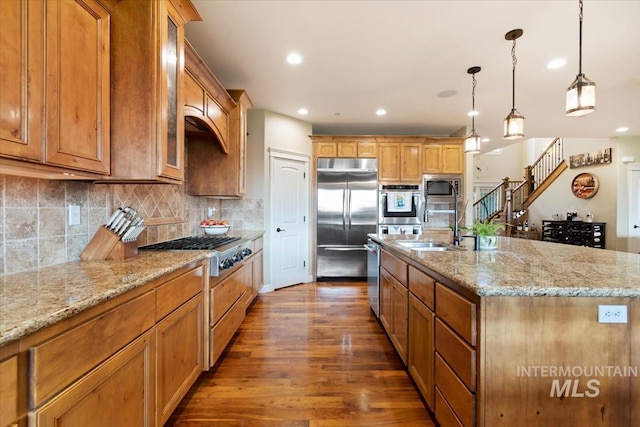 kitchen with tasteful backsplash, an island with sink, hanging light fixtures, built in appliances, and light stone countertops