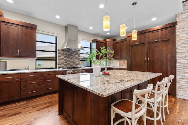 kitchen with decorative backsplash, light wood-style floors, a center island, wall chimney exhaust hood, and hanging light fixtures