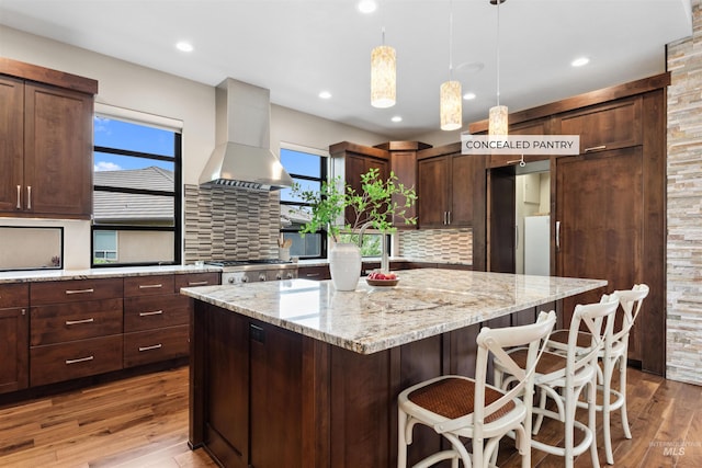 kitchen featuring wood finished floors, a kitchen island, wall chimney range hood, tasteful backsplash, and light stone counters