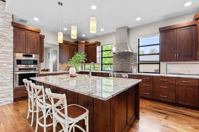kitchen featuring light wood-type flooring, backsplash, a center island with sink, and wall chimney range hood