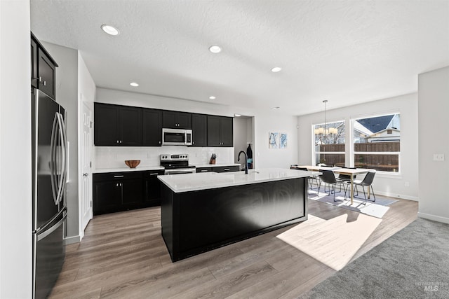 kitchen with decorative light fixtures, decorative backsplash, a kitchen island with sink, light hardwood / wood-style floors, and stainless steel appliances