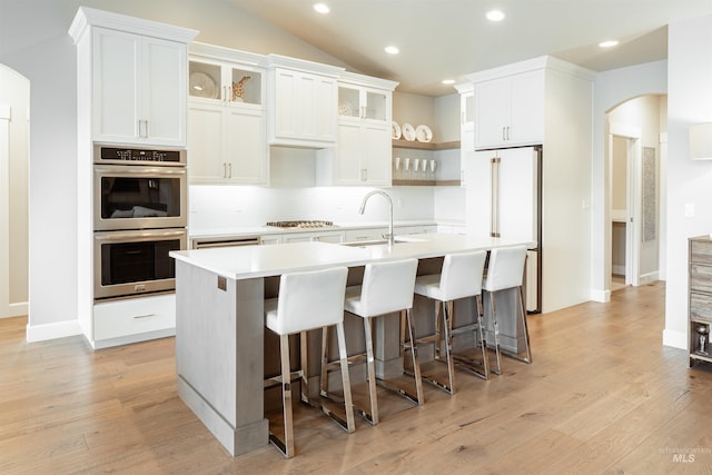 kitchen featuring open shelves, a sink, appliances with stainless steel finishes, arched walkways, and a center island with sink