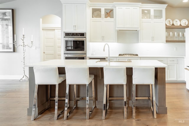 kitchen with a kitchen island with sink, backsplash, light countertops, arched walkways, and appliances with stainless steel finishes