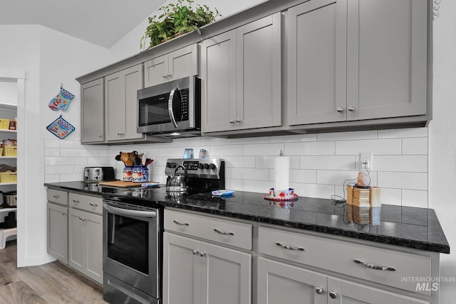 kitchen featuring gray cabinetry, light wood-type flooring, decorative backsplash, dark stone countertops, and appliances with stainless steel finishes