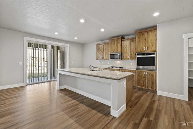 kitchen featuring brown cabinetry, a center island with sink, appliances with stainless steel finishes, dark wood-style floors, and a sink