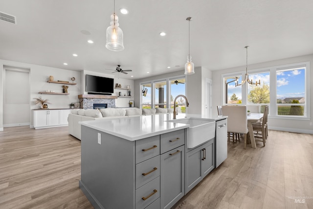 kitchen featuring sink, an island with sink, ceiling fan with notable chandelier, gray cabinets, and a stone fireplace