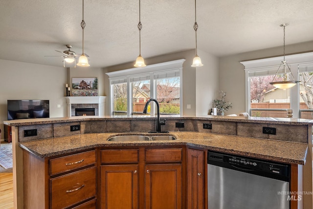 kitchen with a sink, ceiling fan, brown cabinetry, stainless steel dishwasher, and a tile fireplace