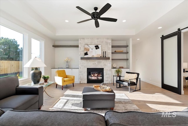 living room with a tray ceiling, a barn door, light wood-style floors, a ceiling fan, and a stone fireplace