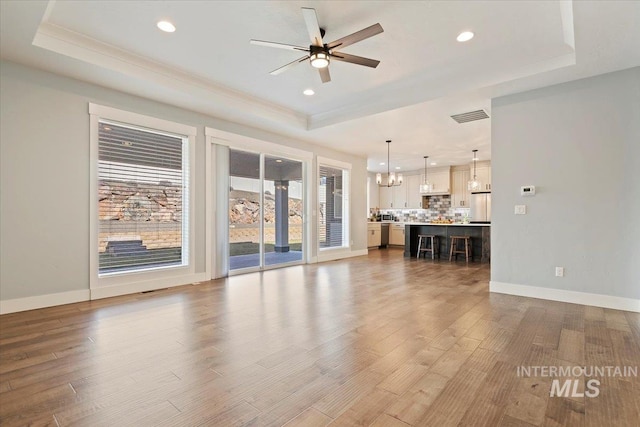 unfurnished living room with ceiling fan with notable chandelier, a raised ceiling, baseboards, and wood finished floors