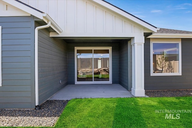 view of exterior entry featuring a yard, a patio area, board and batten siding, and roof with shingles