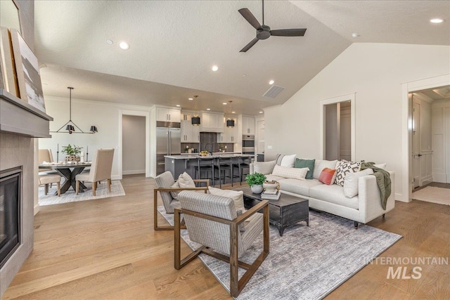 living room with ceiling fan with notable chandelier, vaulted ceiling, light wood-type flooring, a textured ceiling, and a tiled fireplace