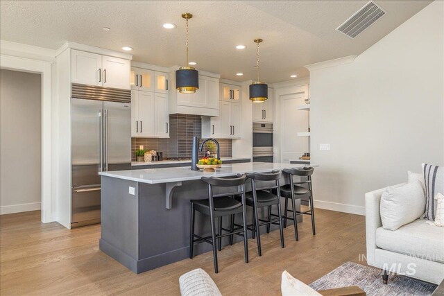 kitchen featuring a kitchen island with sink, white cabinets, decorative backsplash, decorative light fixtures, and stainless steel appliances