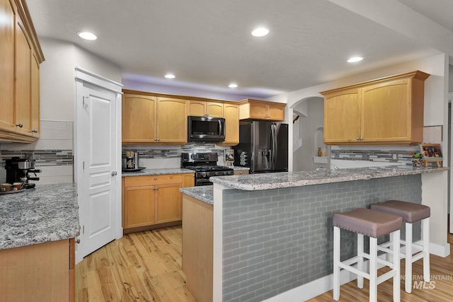 kitchen with arched walkways, light wood-style flooring, appliances with stainless steel finishes, light stone countertops, and a kitchen breakfast bar