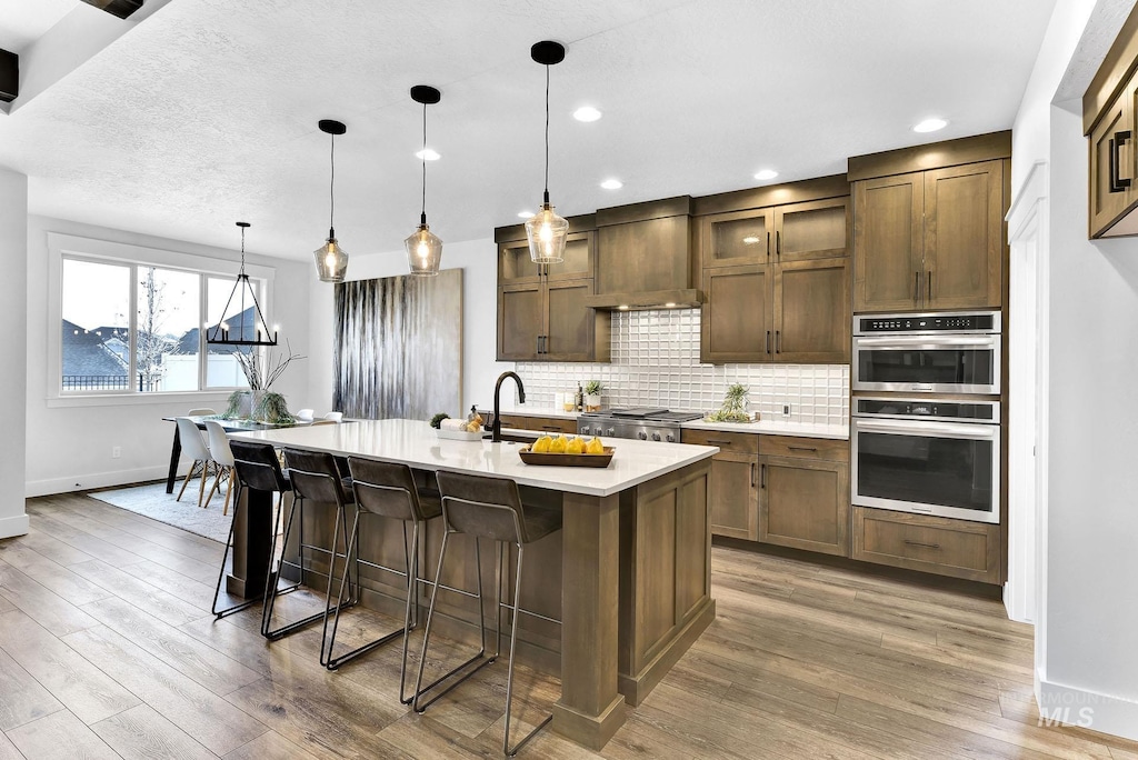 kitchen featuring premium range hood, a center island with sink, double oven, decorative light fixtures, and a breakfast bar area