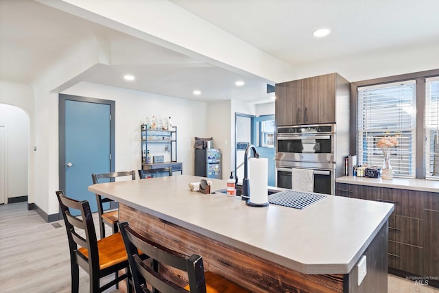 kitchen featuring a breakfast bar, a kitchen island with sink, light hardwood / wood-style floors, dark brown cabinetry, and double oven