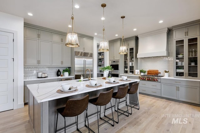 kitchen featuring a center island with sink, decorative backsplash, custom exhaust hood, gray cabinetry, and appliances with stainless steel finishes