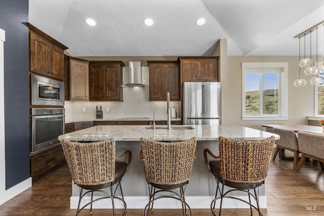 kitchen with dark wood-style floors, an island with sink, stainless steel appliances, a sink, and wall chimney exhaust hood