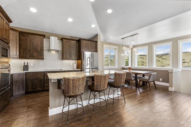 kitchen featuring lofted ceiling, an island with sink, dark wood-style floors, stainless steel appliances, and wall chimney exhaust hood
