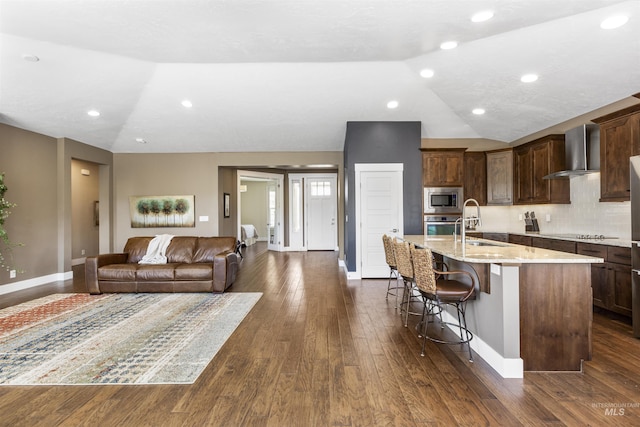 kitchen with an island with sink, stainless steel appliances, vaulted ceiling, a sink, and wall chimney exhaust hood