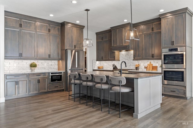 kitchen with pendant lighting, appliances with stainless steel finishes, dark wood-type flooring, a kitchen breakfast bar, and a center island with sink