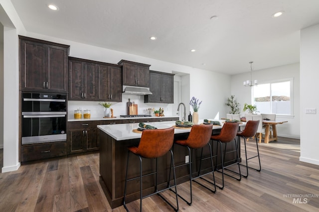 kitchen featuring stainless steel gas stovetop, under cabinet range hood, double wall oven, light countertops, and a kitchen bar